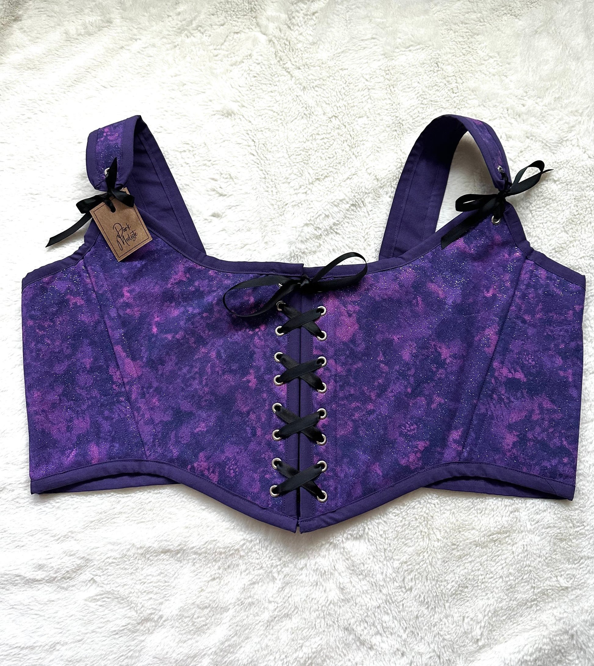 Renaissance bodice in purple and pink celestial print. Purple bias border around edges, black ribbon down center with silver grommets. Ribbon connects the straps to the bodice with a "Dark Modiste" tag on top left strap. Bodice is laying on white fluffy blanket.