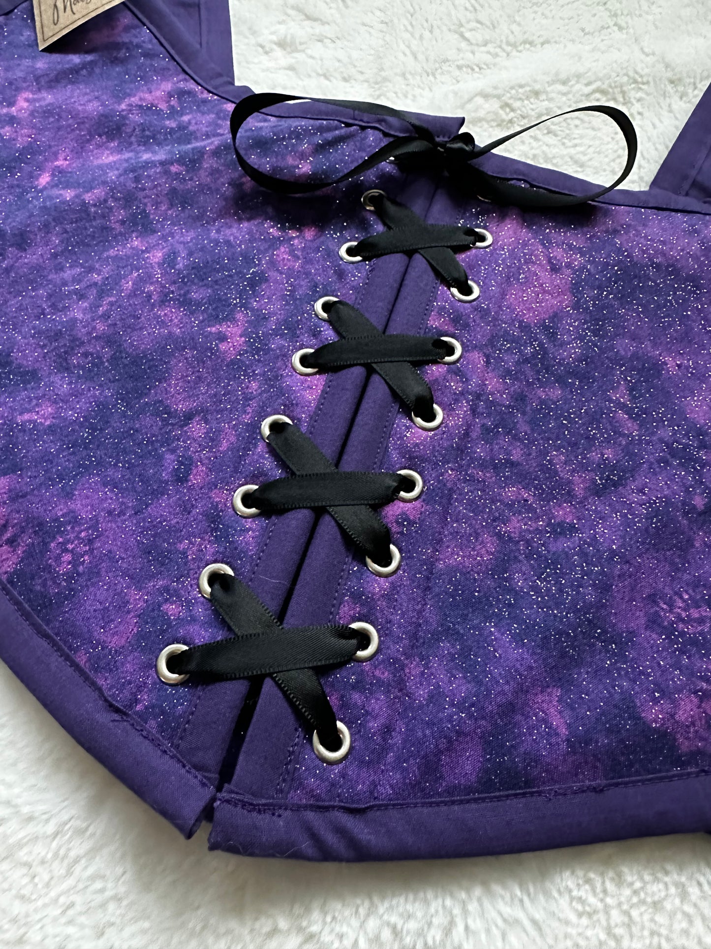 Close up of renaissance bodice in purple and pink celestial print. Purple bias border around edges, black ribbon down center with silver grommets. Bodice is laying on white fluffy blanket.