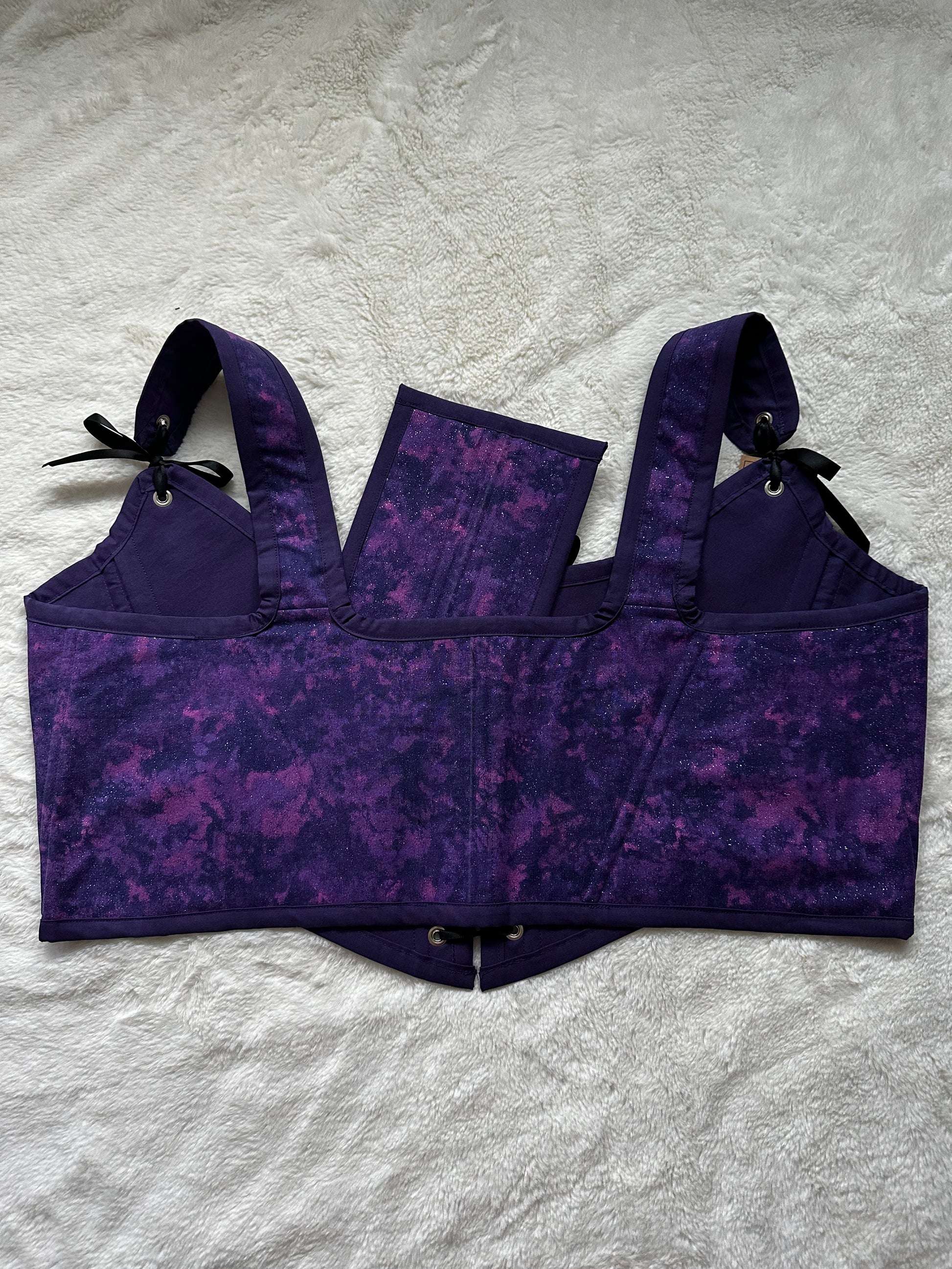 Back view of renaissance bodice in purple and pink celestial print. Purple bias border around edges. Black ribbon connects the straps to the bodice. Bodice is laying on white fluffy blanket.