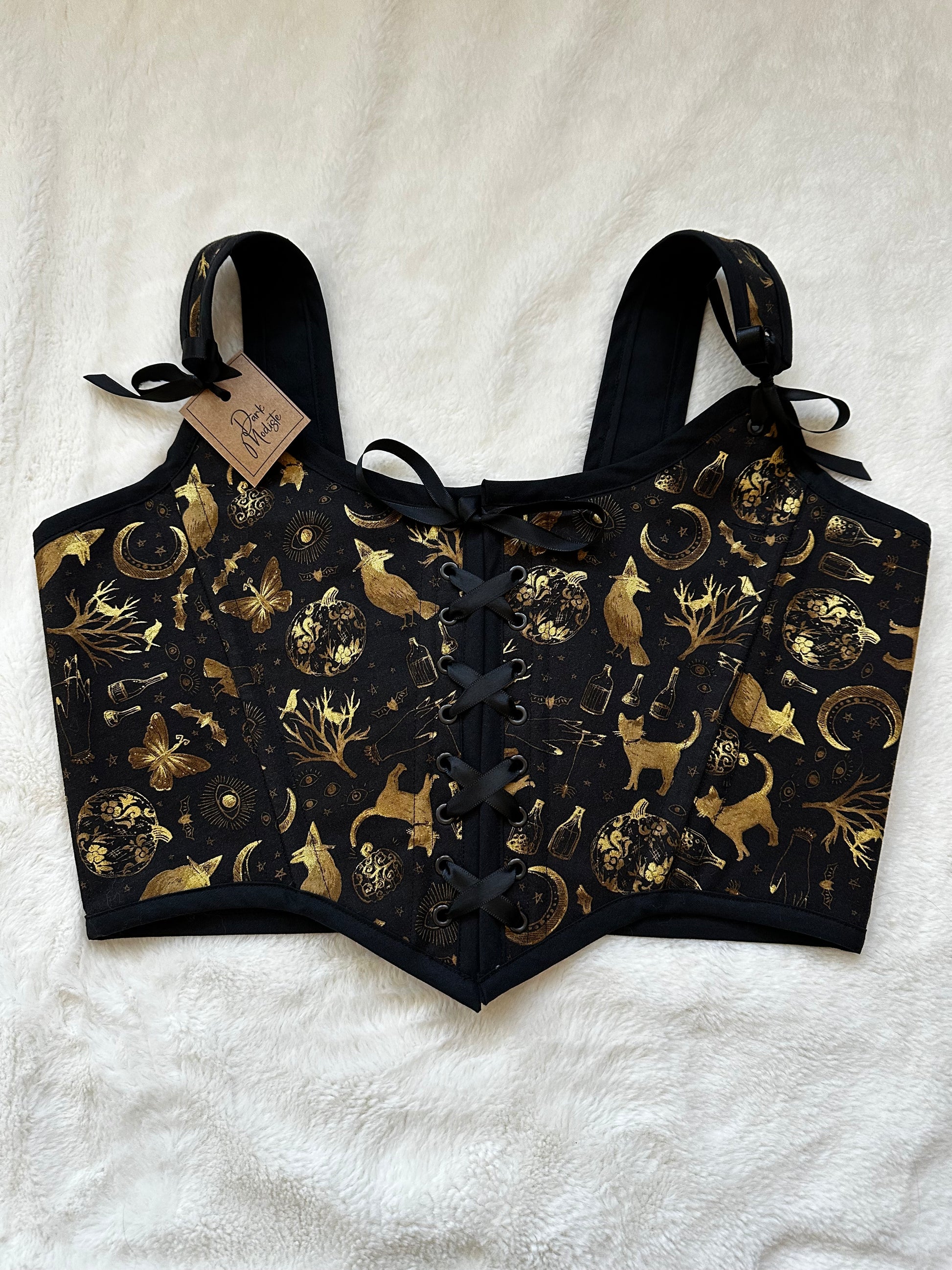 Renaissance bodice in black fabric with gold patterned print. Pattern contains cats, trees, moons, bottles, and moths in gold. Black ribbon is tied in the middle and in the straps. There is a "Dark Modiste" tag in the left strap. Bodice is on a white fuzzy blanket.