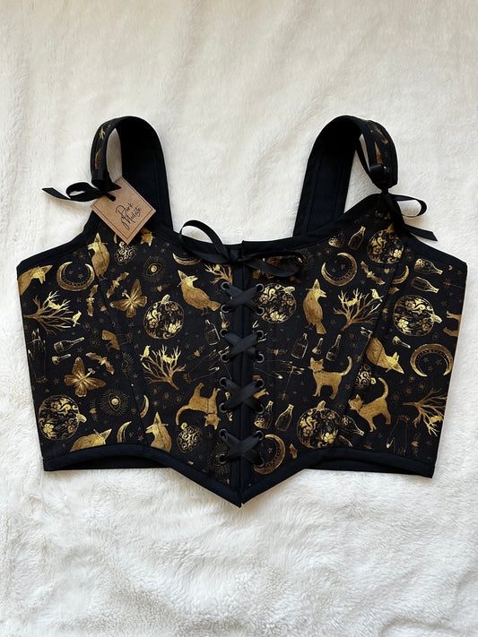 Renaissance bodice in black fabric with gold patterned print. Pattern contains cats, trees, moons, bottles, and moths in gold. Black ribbon is tied in the middle and in the straps. There is a "Dark Modiste" tag in the left strap. Bodice is on a white fuzzy blanket.
