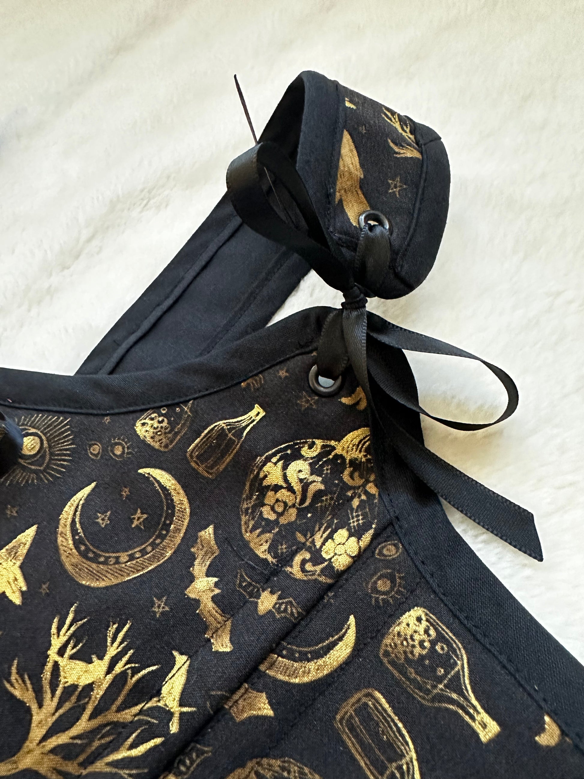 Top right strap and front of renaissance bodice in black fabric with gold patterned print. Pattern contains trees, moons, bottles, and bats in gold. Strap is tied to bodice with black ribbon. Bodice is on a white fuzzy blanket.