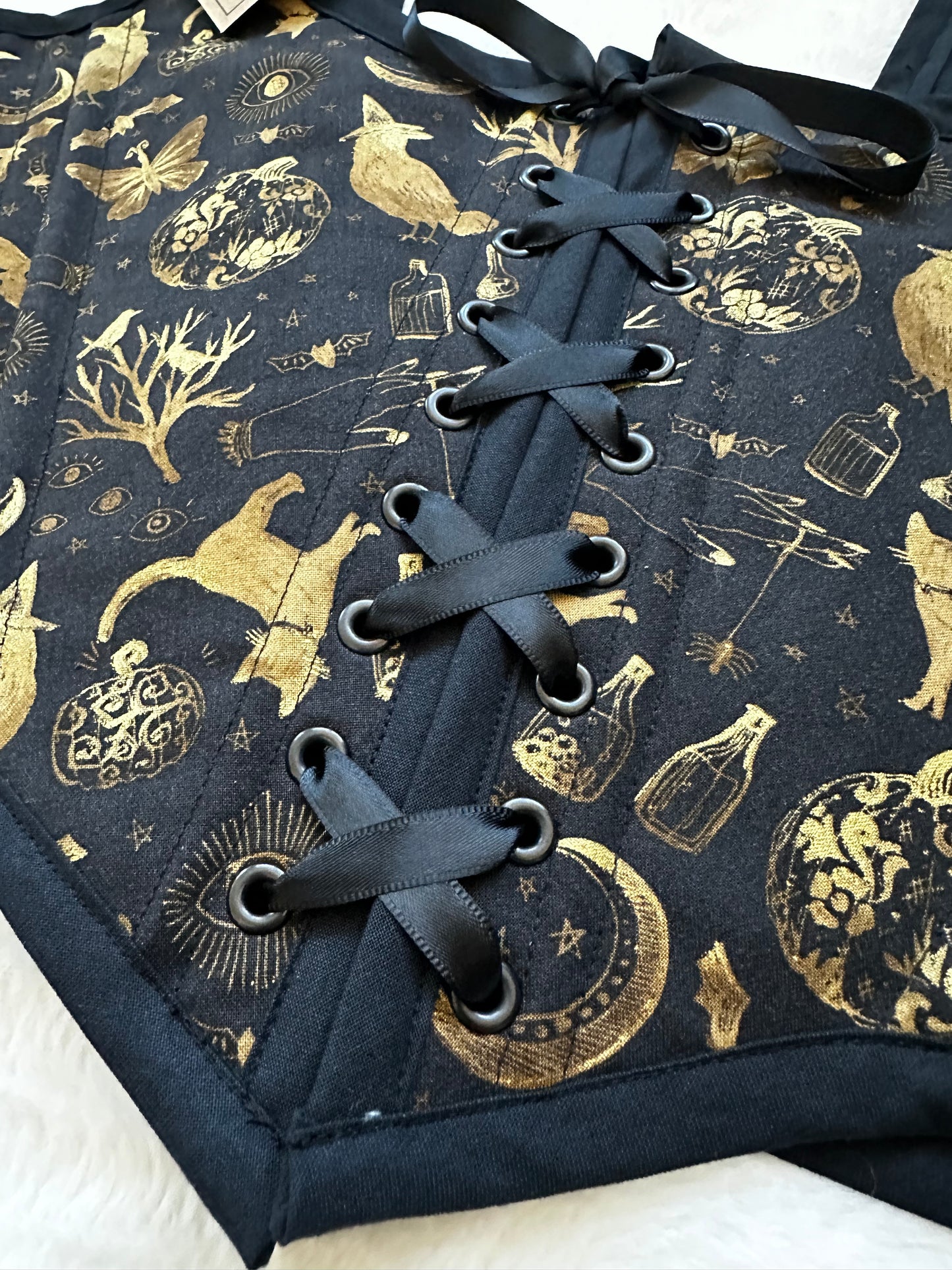 Close up of renaissance bodice in black fabric with gold patterned print. Pattern contains cats, trees, moons, bottles, and moths in gold. Black ribbon is tied in the middle. Bodice is on a white fuzzy blanket.