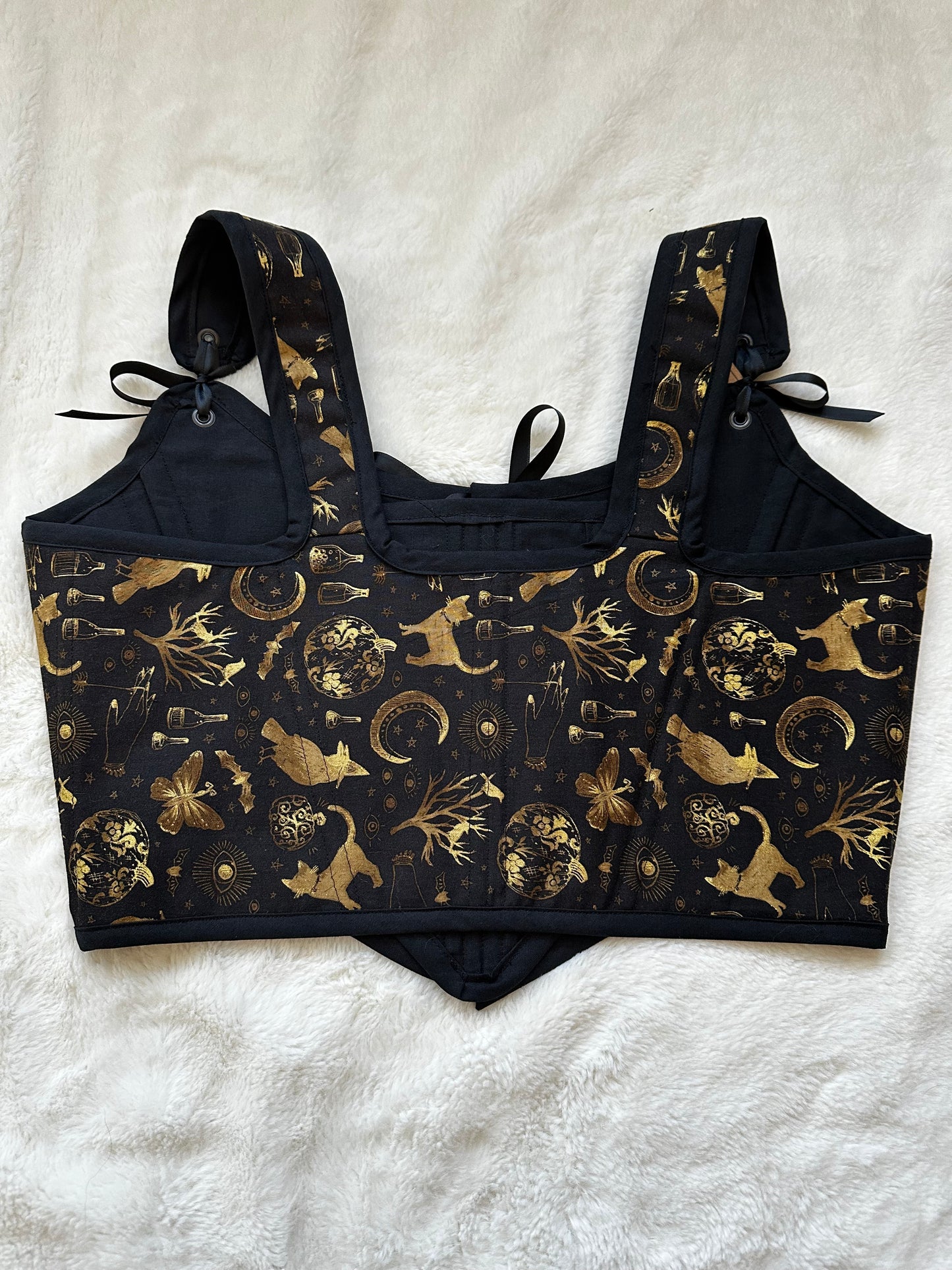 Back view of renaissance bodice in black fabric with gold patterned print. Pattern contains cats, trees, moons, bottles, and moths in gold. Black ribbon connects the bodice to the straps. Bodice is on a white fuzzy blanket.