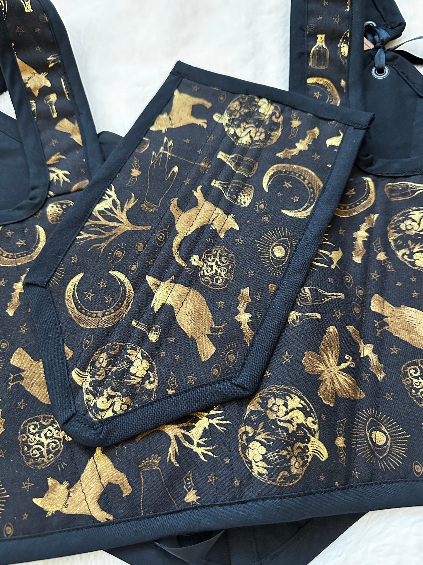Back view of renaissance bodice in black fabric with gold patterned print. Pattern contains cats, trees, moons, bottles, and moths in gold. Also shown is the stomacher. Bodice is on a white fuzzy blanket.